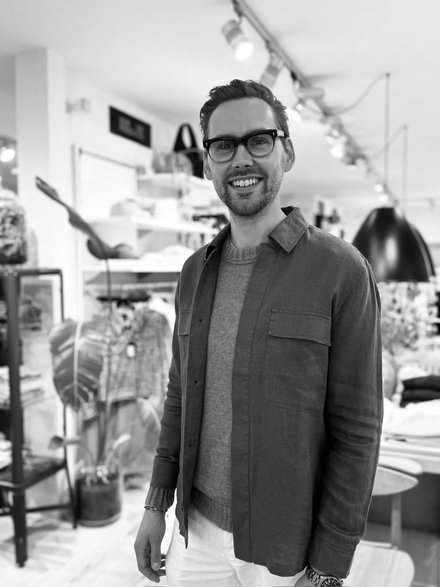 Mand med briller, grå sweater, åben skjorte og hvide bukser i butik.