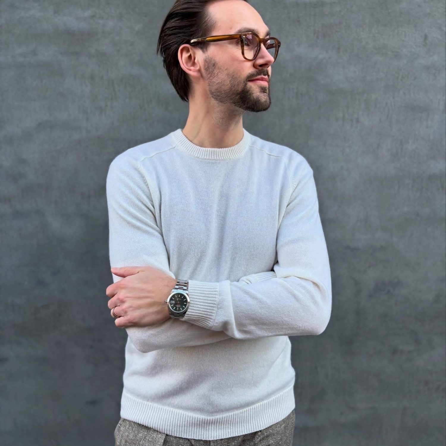 Mand med briller og hvid striktrøje står foran grå mur, kigger til siden.